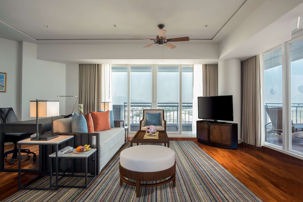 Ruime hotelkamer met groot raam, plafondventilator, flatscreen televisie, bank, salontafel, klein bijzettafeltje en een rond hocker.