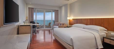 A modern hotel room with a large bed, a desk, and a view of the sea through the glass doors.