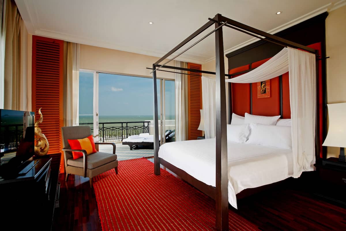 Een bed met vier poten, voorzien van witte lakens, een rood tapijt, een televisie en een stoel met oranje kussens in een kamer met een rode accentmuur en uitzicht op de oceaan.