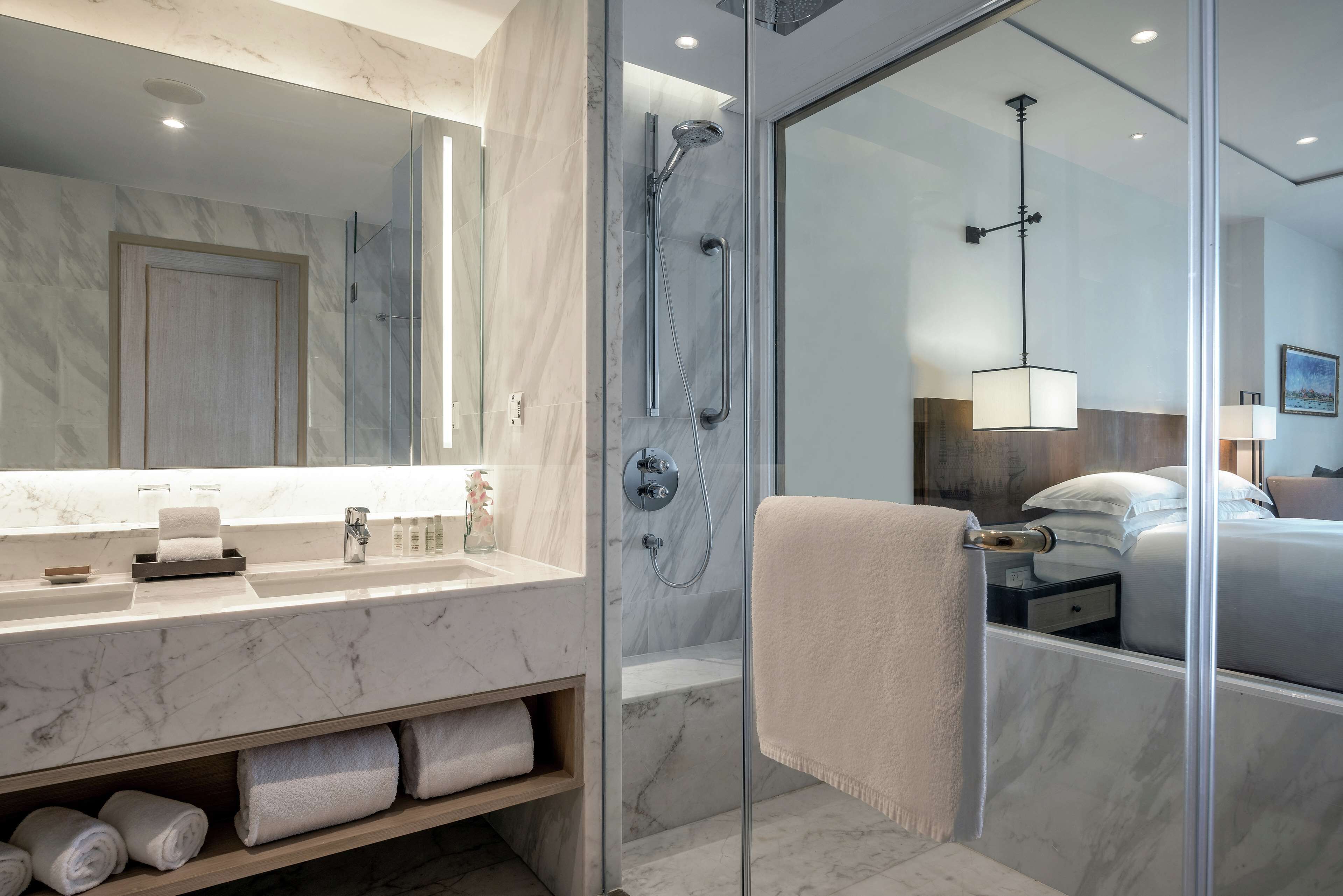 A modern bathroom with a marble vanity, a glass shower enclosure, and a visible bedroom with a bed and nightstand.