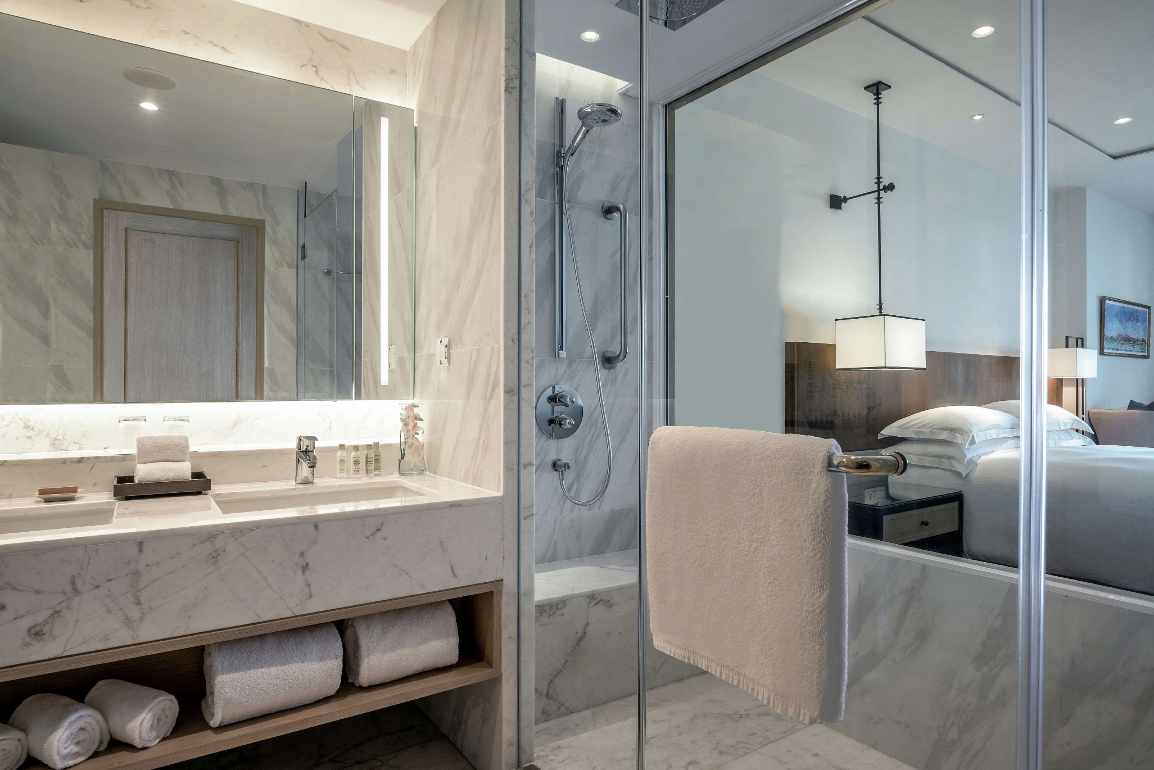 Un baño moderno con tocador de mármol, cabina de ducha de vidrio y una habitación visible con cama y mesita de noche.