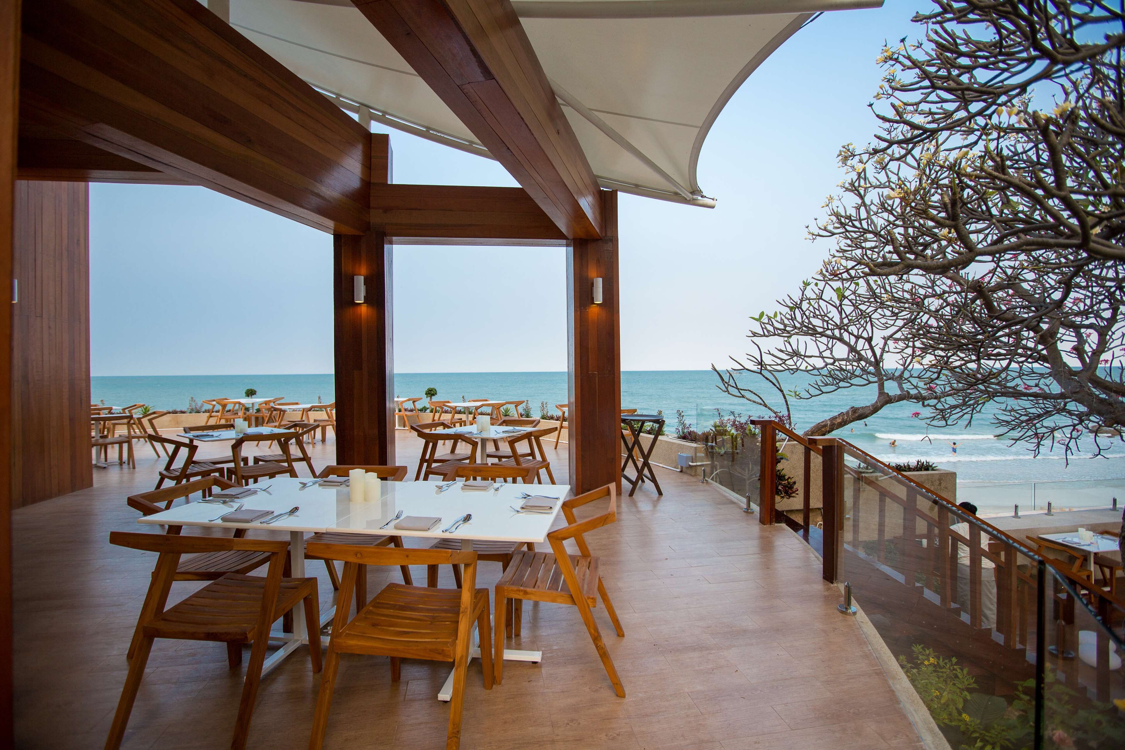 Un espace de restauration en plein air, avec des tables et des chaises en bois, offrant une vue sur la plage et l’océan.