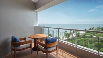 A balcony with two wooden chairs and a small round table, overlooking a beach and lush greenery.