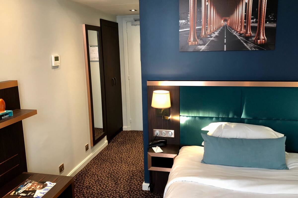 Een hotelkamer met een bed, nachtkastje, lamp, spiegel, kledingkast en een schilderij van een brug.