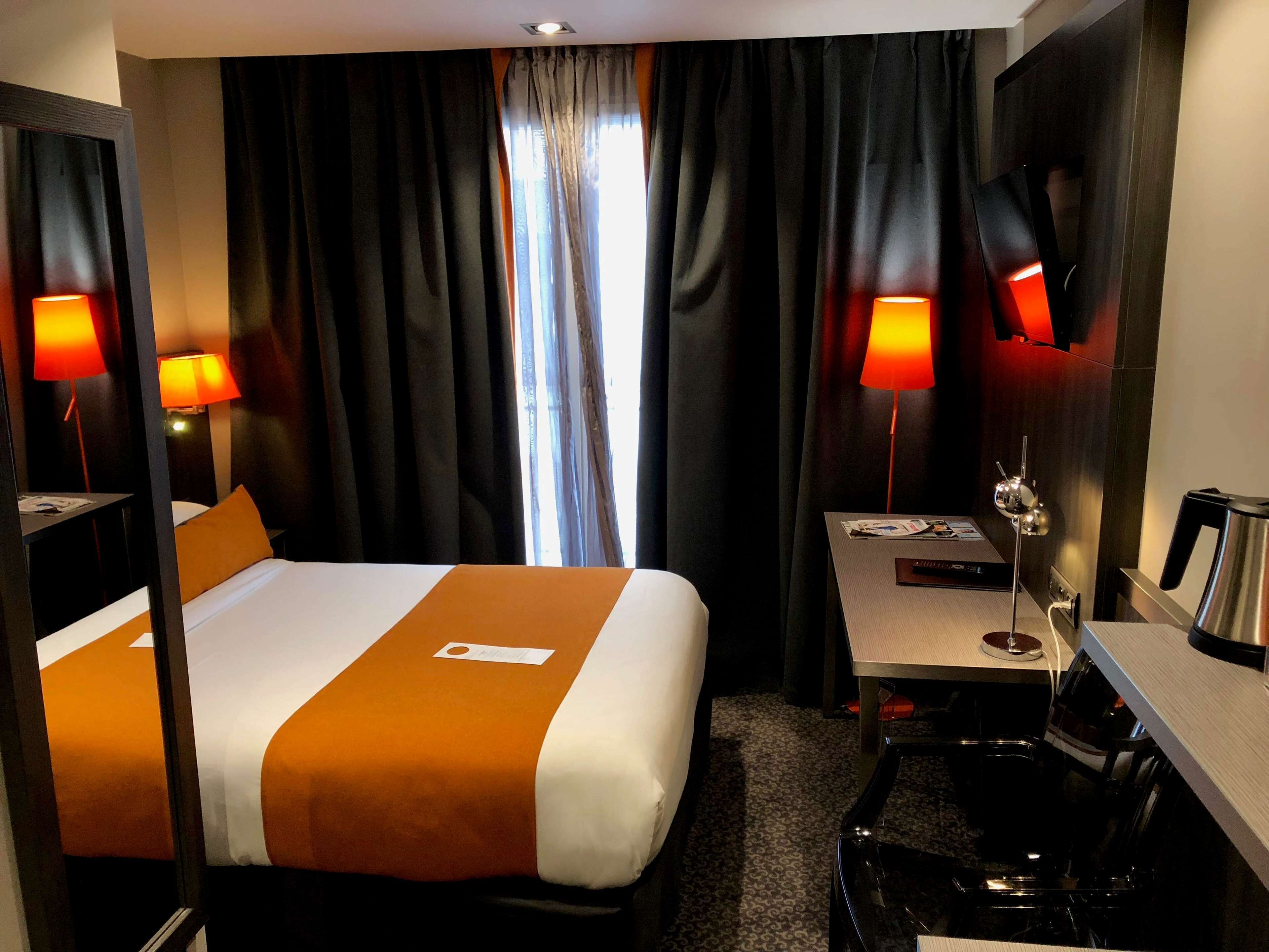 A hotel room with a bed, two bedside lamps, a desk with a chair, a coffee maker, and a mirror.