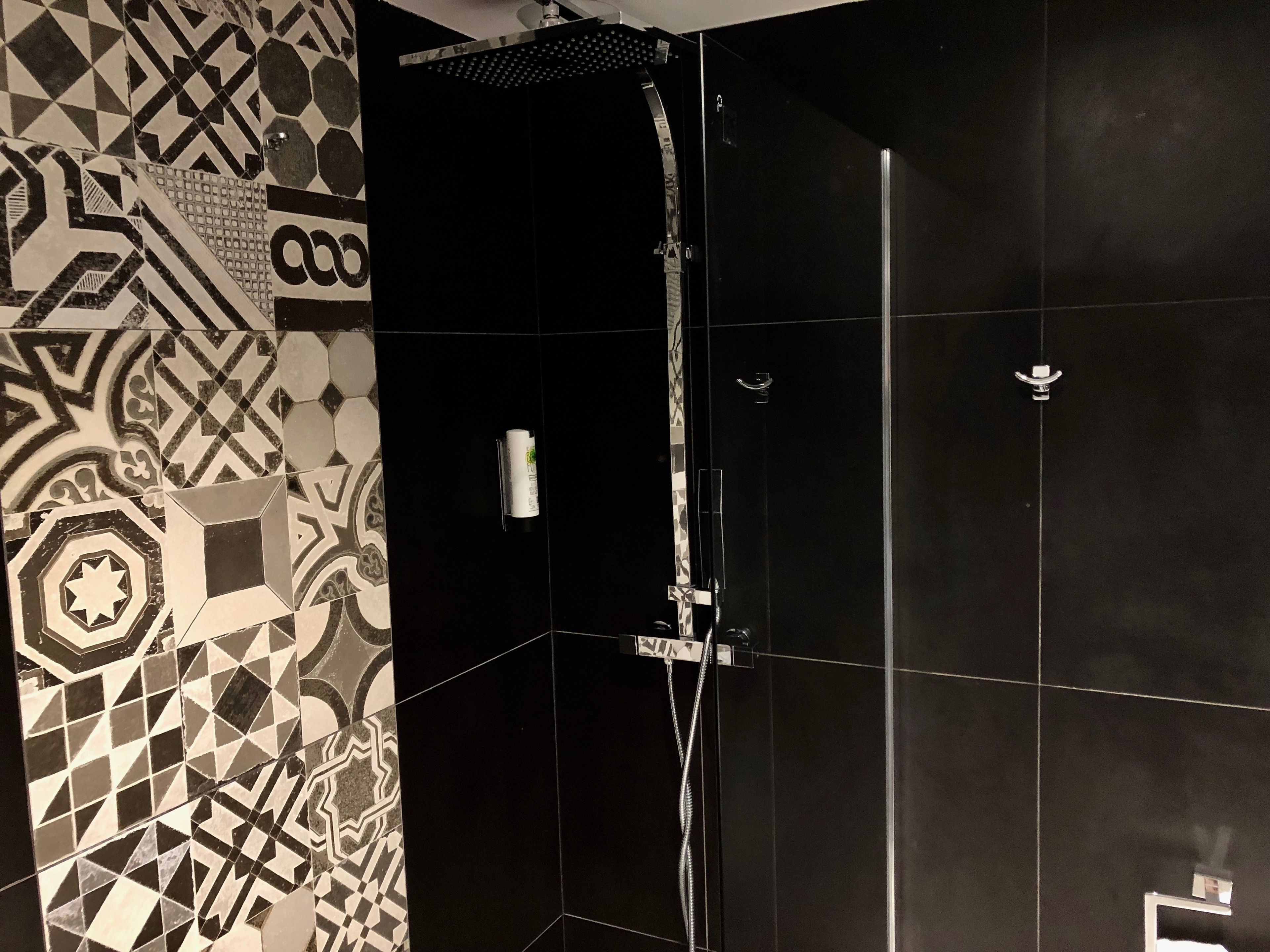 Une salle de bain moderne avec un motif géométrique sur les carreaux du mur de gauche, une douche dont le sol est recouvert de carreaux noirs et un sèche-mains visible.
