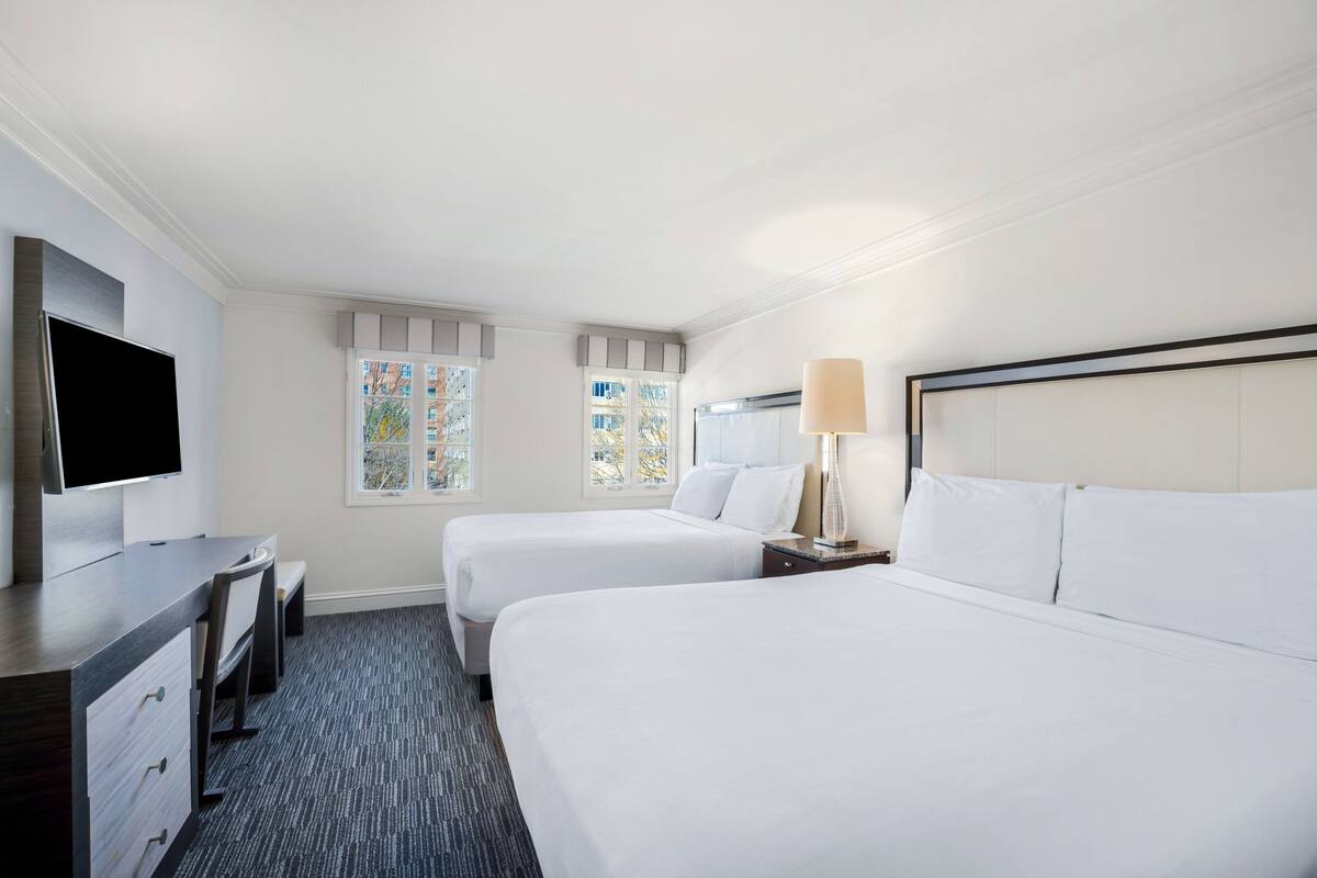 A hotel room with two beds, a desk, a TV, and a window with a view of buildings.
