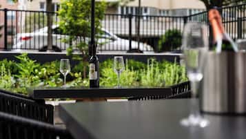 Una mesa con una botella de vino y dos copas, ubicada al aire libre con follaje y una valla de fondo.