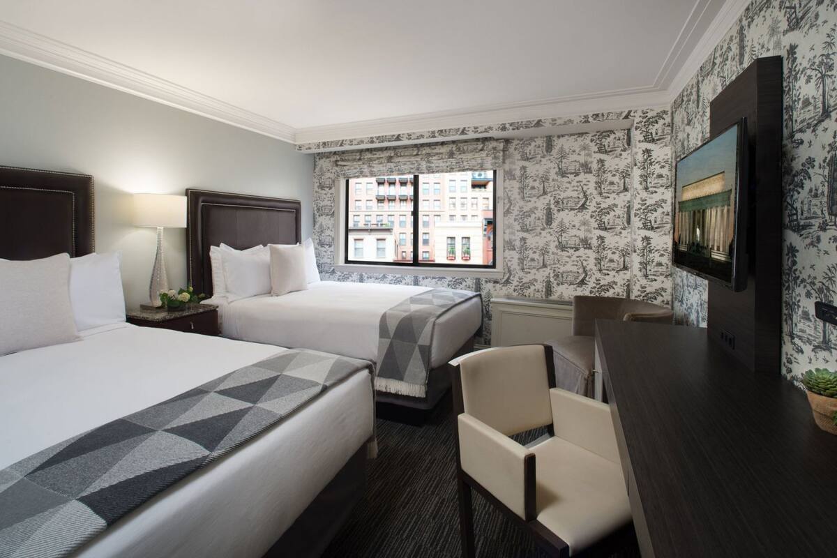 A hotel room with two beds, a desk, a chair, a TV, and a window with a city view.