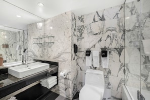 Features a sleek sink, white vanity, and marble walls with a glass shelf for toiletries.