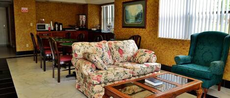 A living room with a floral sofa, a green armchair, a wooden coffee table, and a dining area with a microwave and a waste bin.