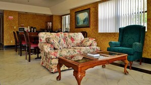 A hotel lobby with a floral sofa, a green armchair, a wooden coffee table, a dining area with chairs and a table, and a painting on the wall. - Executive Inn Opp (Opp)