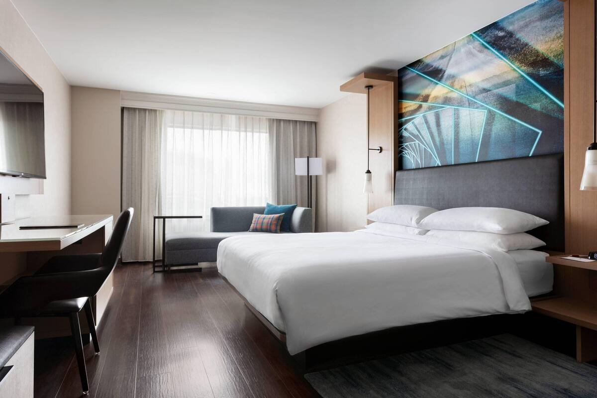 A modern hotel room with a large bed, a desk, a chair, and a large window with curtains.