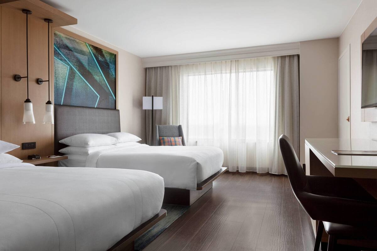 A hotel room with two beds, a desk, a chair, a large window with curtains, and a painting on the wall.