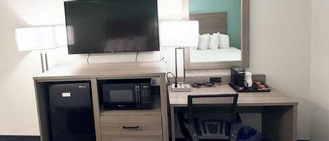 A hotel room with a desk, a television, a microwave, a chair, a trash bin, and a coffee maker.