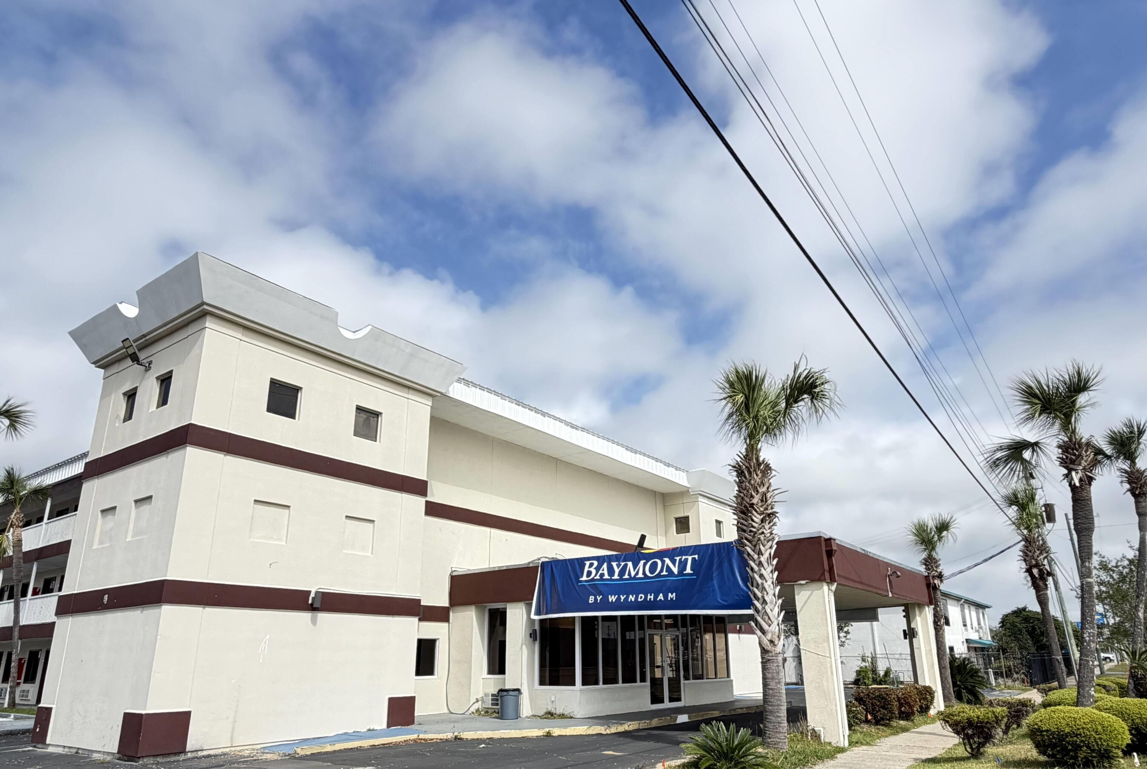 Un hotel con fachada blanca, un toldo azul con el rótulo 'BAYMONT BY WYNDHAM' y un estacionamiento con palmeras.