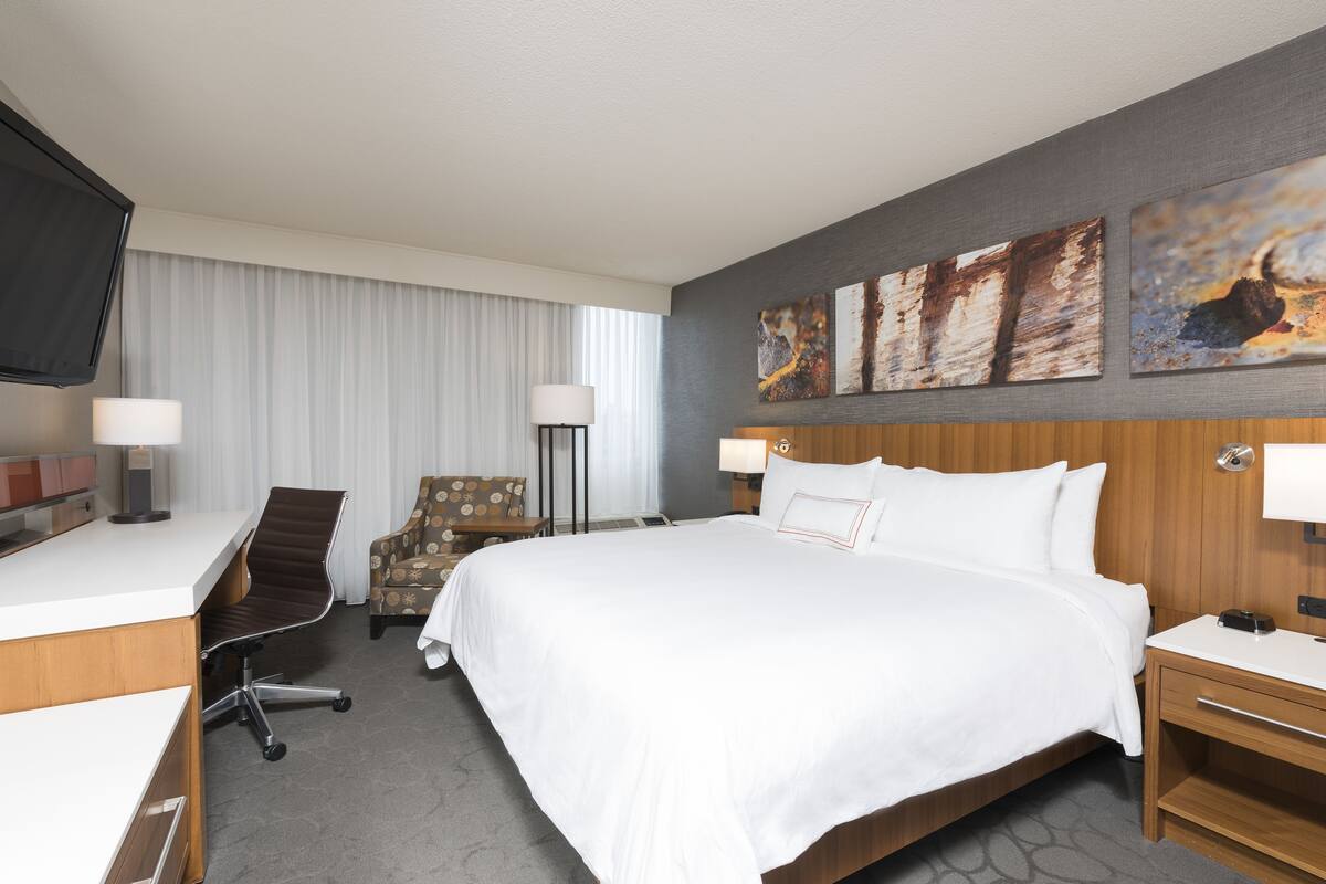 A hotel room with a bed, desk, chair, and artwork on the wall.