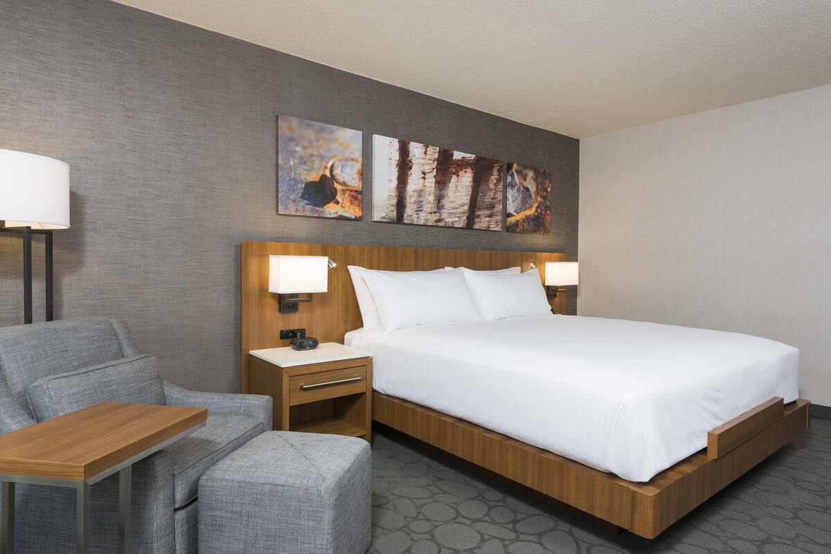 A hotel room with a large bed, a bedside table with a lamp, a chair, and artwork on the wall.