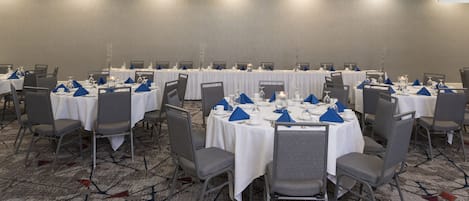 Sala de conferência com mesas redondas, cadeiras cinzas, toalhas de mesa brancas e suportes para guardanapos azuis.