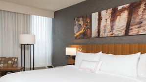 A hotel room with a large bed, a nightstand with a lamp, a chair, and artwork on the wall. - The Center Hotel Grand Rapids Airport (Grand Rapids)