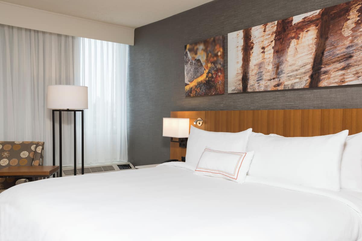 A hotel room with a large bed, a nightstand with a lamp, a chair, and artwork on the wall.