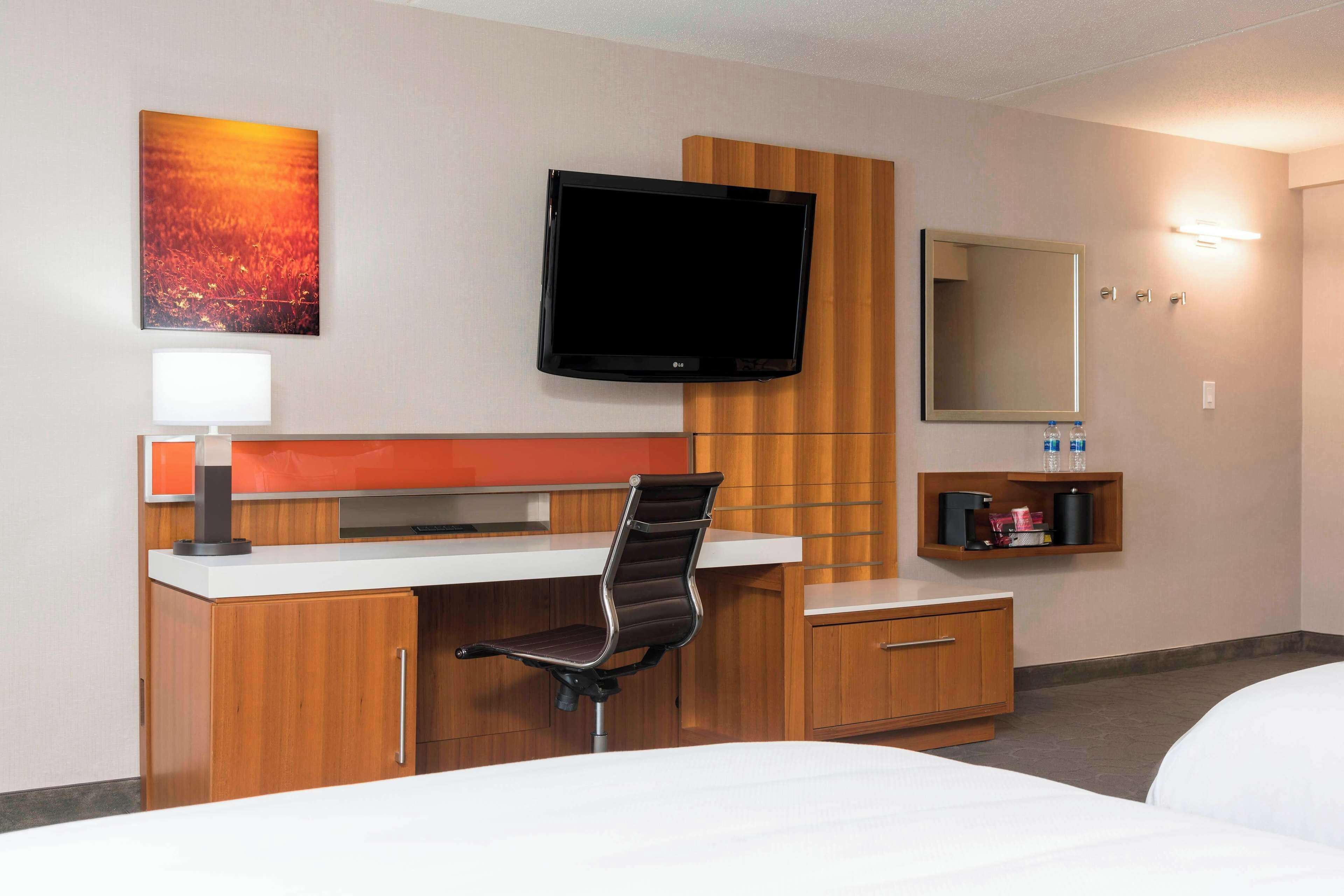 A hotel room with a bed, a desk with a chair, a flat-screen TV, a mirror, and a shelf with toiletries.