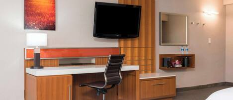 A hotel room with a bed, a desk with a chair, a flat-screen TV, a mirror, and a shelf with toiletries.