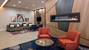 A modern lounge area with a sofa set, two red armchairs, a glass coffee table, and a wall-mounted fireplace. - The Center Hotel Grand Rapids Airport (Grand Rapids)