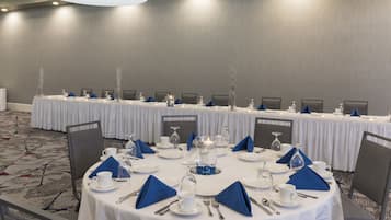 Un salón de conferencias con mesas redondas dispuestas para una reunión, equipado con manteles blancos, servilletas azules y cristalería.