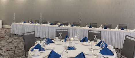Un salón de conferencias con mesas redondas dispuestas para una reunión, equipado con manteles blancos, servilletas azules y cristalería.