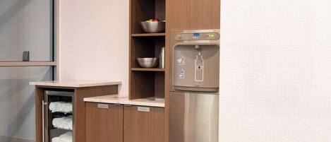 A compact kitchenette with a built-in refrigerator, a water dispenser, and a glass-fronted storage unit with towels.