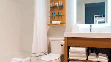Et badeværelse med toilet, badeforhæng, vask med spejl og hylder med toiletartikler og håndklæder.