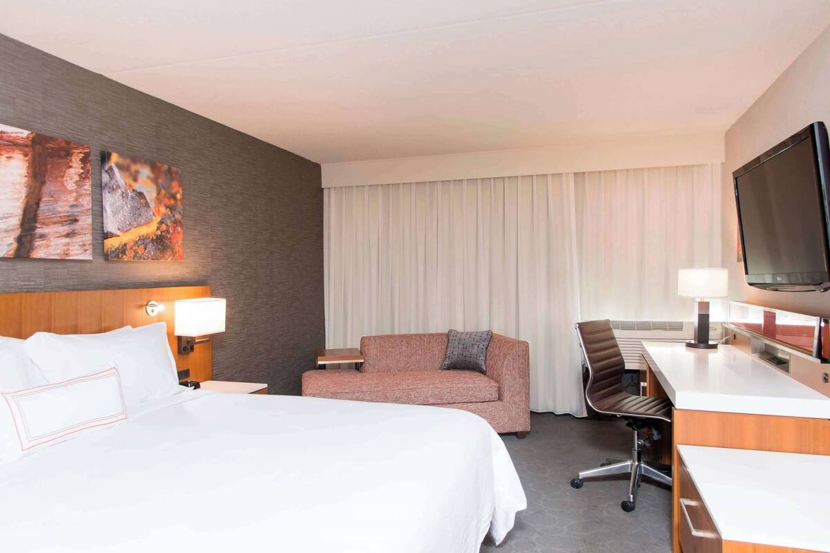 A hotel room with a bed, a sofa, a desk, a chair, a TV, and artwork on the wall.