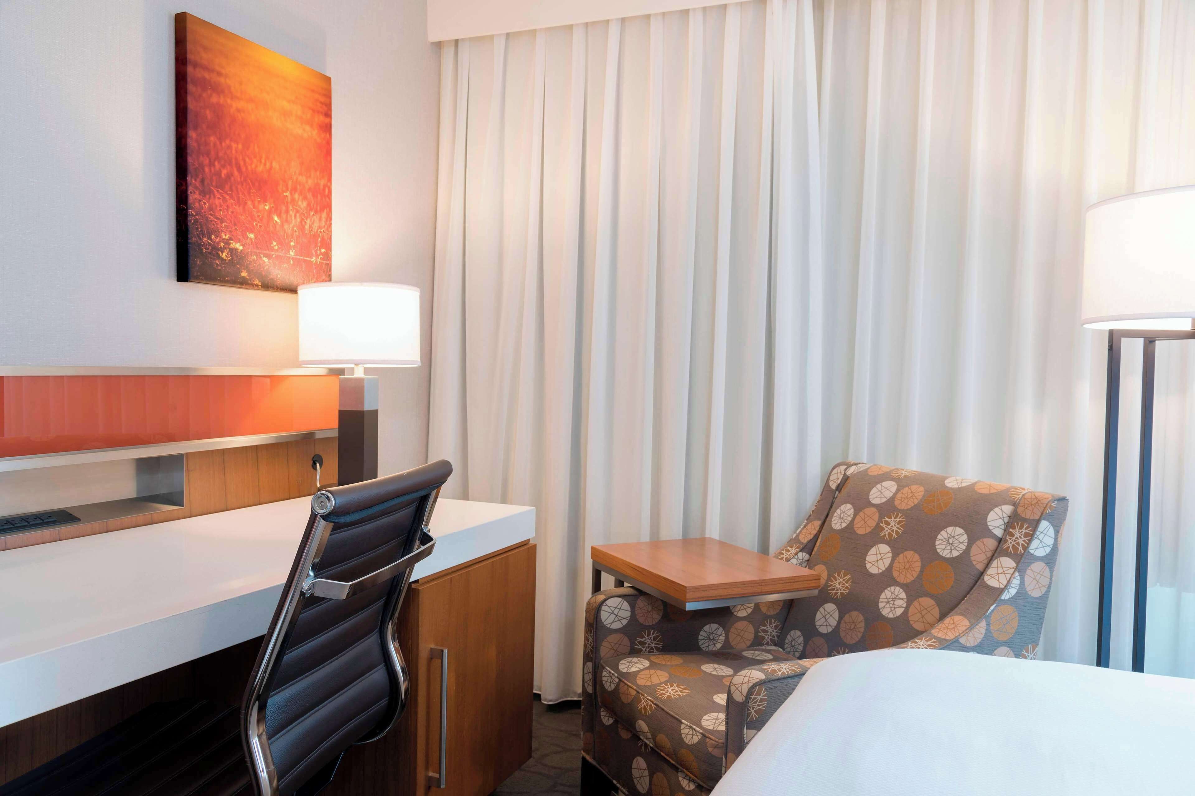 A hotel room with a bed, a desk, a chair, a small table, a lamp, and a window with curtains.