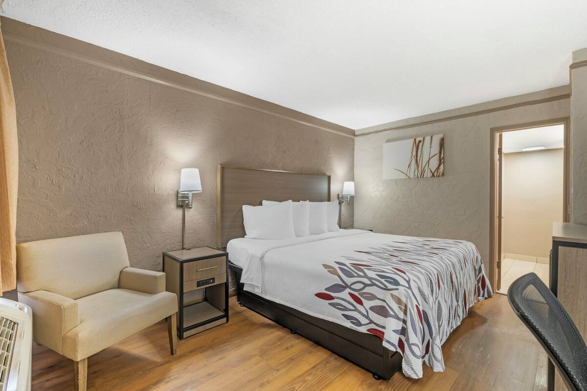 Une chambre d’hôtel comprenant un lit, une table de chevet, une lampe, une chaise et un mur orné d’un tableau.