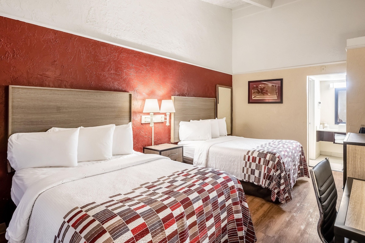 A hotel room with two beds, a nightstand with a lamp, a painting on the wall, and a view of the bathroom.