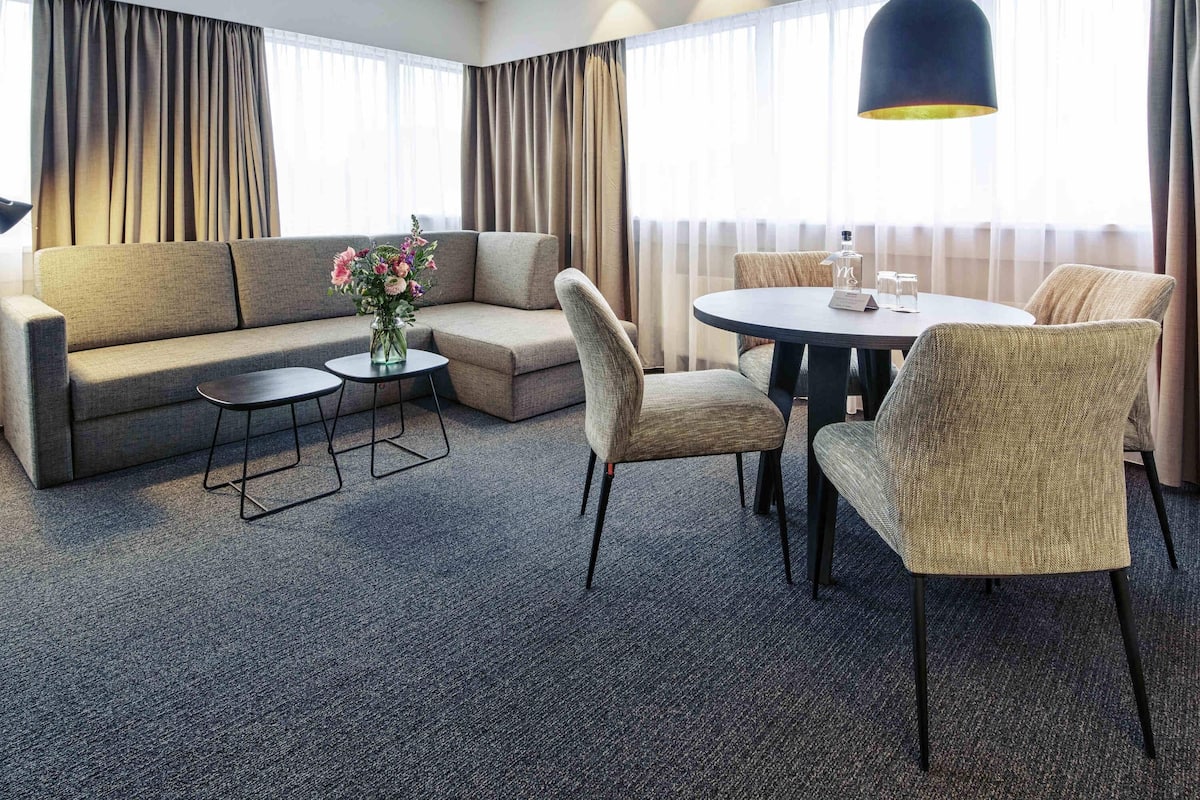 Hotelkamer met een bank, twee fauteuils, een kleine tafel, een ronde eettafel met stoelen, een hanglamp en een bloemenvaas.