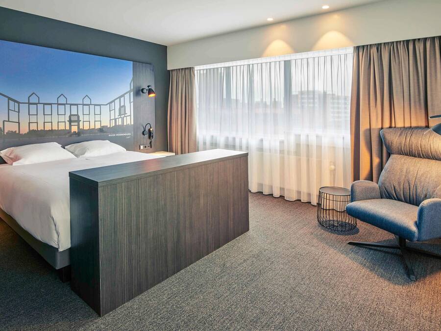 A hotel room with a bed, a desk, a chair, and a large window with curtains.