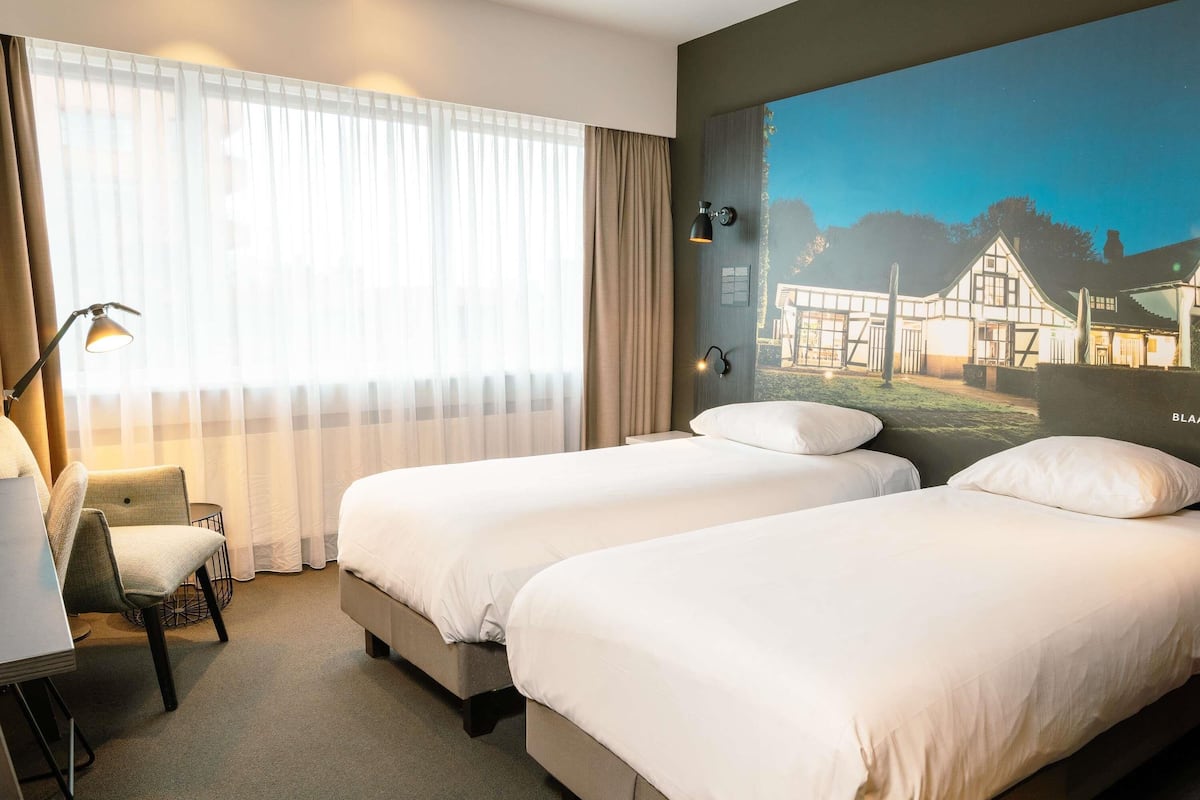 Een hotelkamer met twee bedden, een bureau, een stoel en een groot raam met dunne gordijnen.