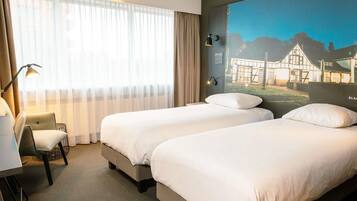 Une chambre d’hôtel avec deux lits, un bureau, une chaise et une grande fenêtre avec des rideaux légers.