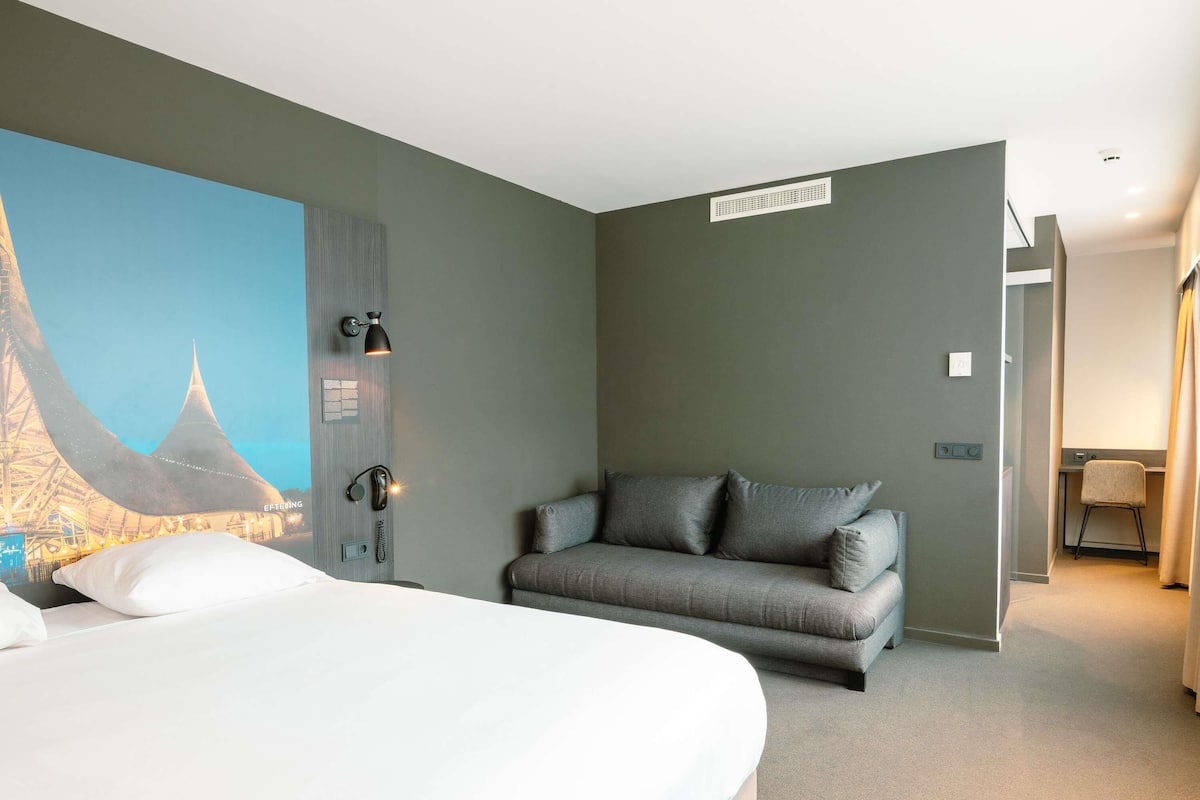 Een hotelkamer met een bed, een grijze bank, een telefoon aan de muur en een schilderij van een brug.
