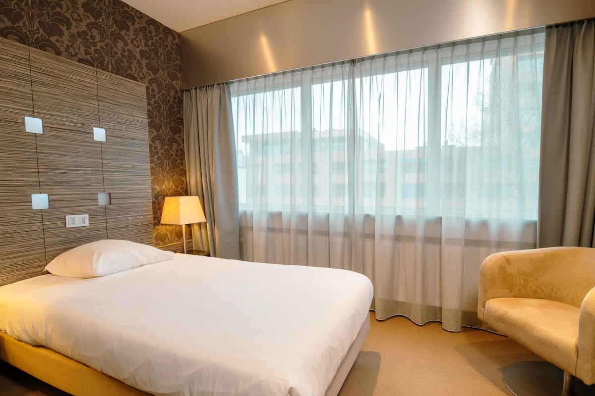 Een hotelkamer met een groot bed, een stoel, een lamp en een raam met gordijnen.