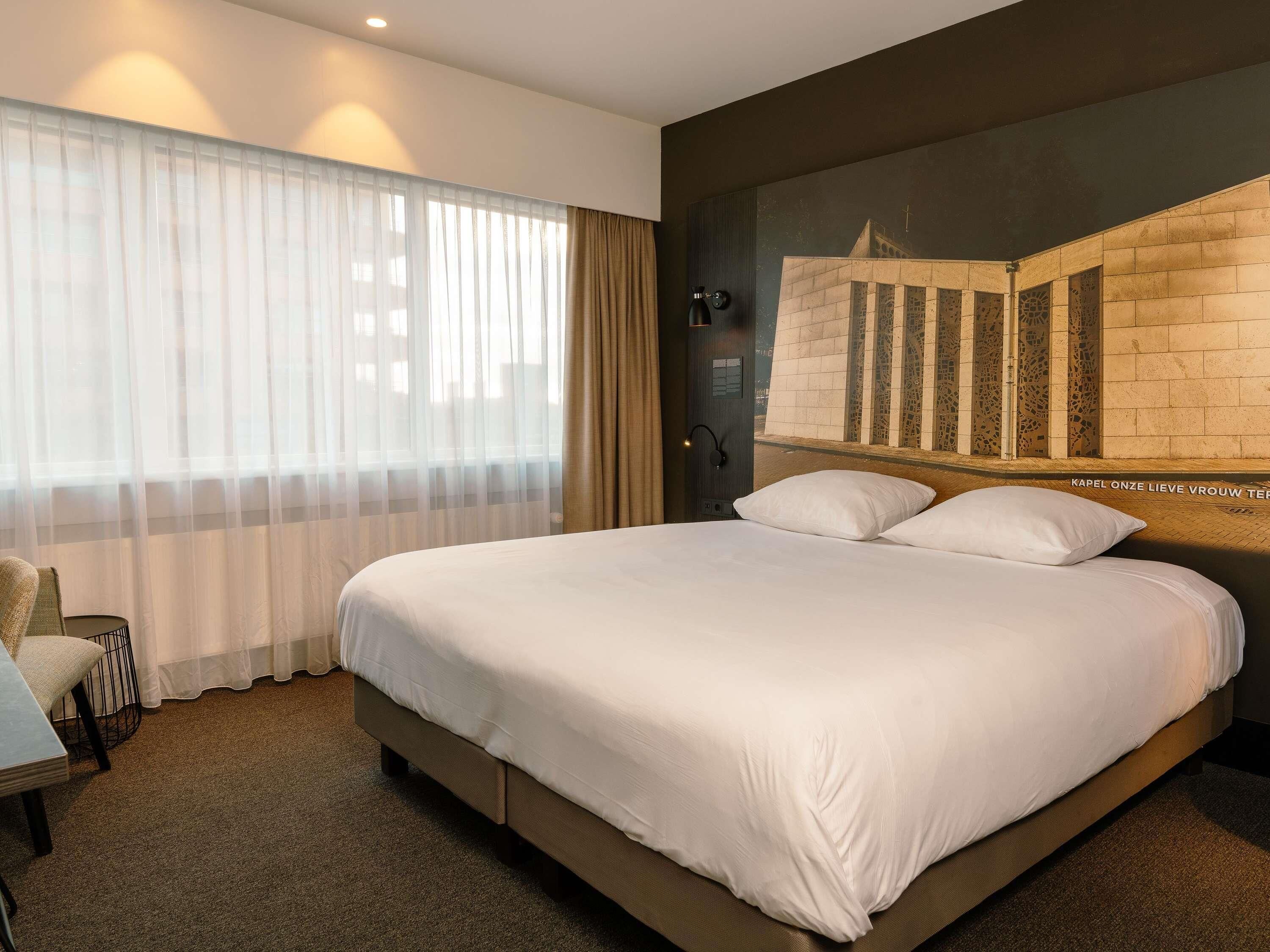 A hotel room with a large bed, a chair, a window with sheer curtains, and a wall-mounted light.
