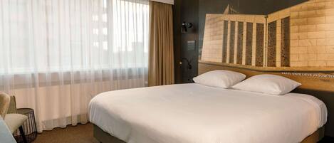 A hotel room with a large bed, a chair, a window with sheer curtains, and a wall-mounted light.