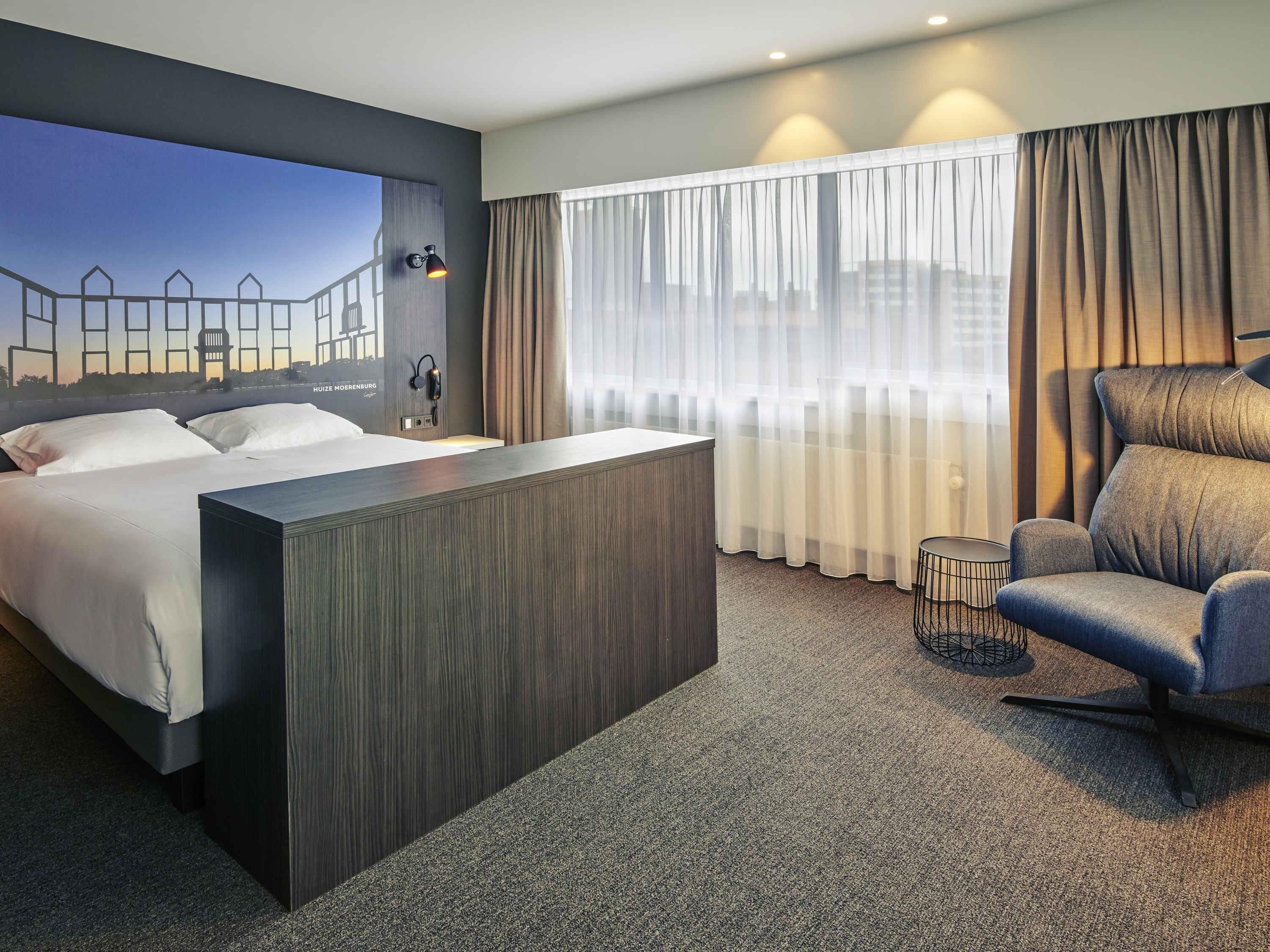 Una habitación de hotel con cama, escritorio, silla y un amplio ventanal con cortinas.