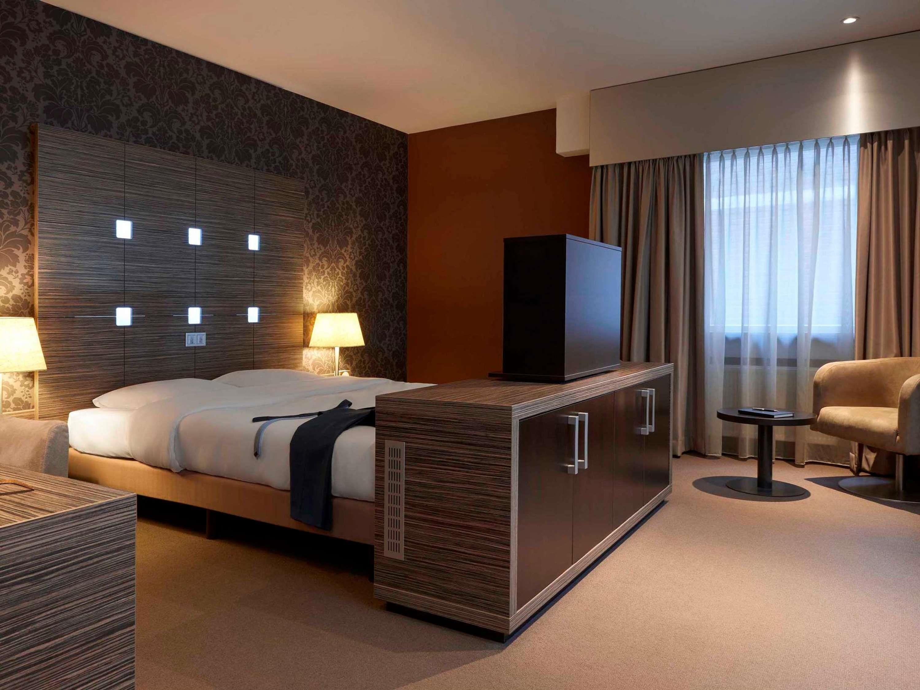 Ein Hotelzimmer mit Bett, Nachttischlampen, einem Fernseher auf einem Schrank, einem Sessel und einem Fenster mit Vorhängen.