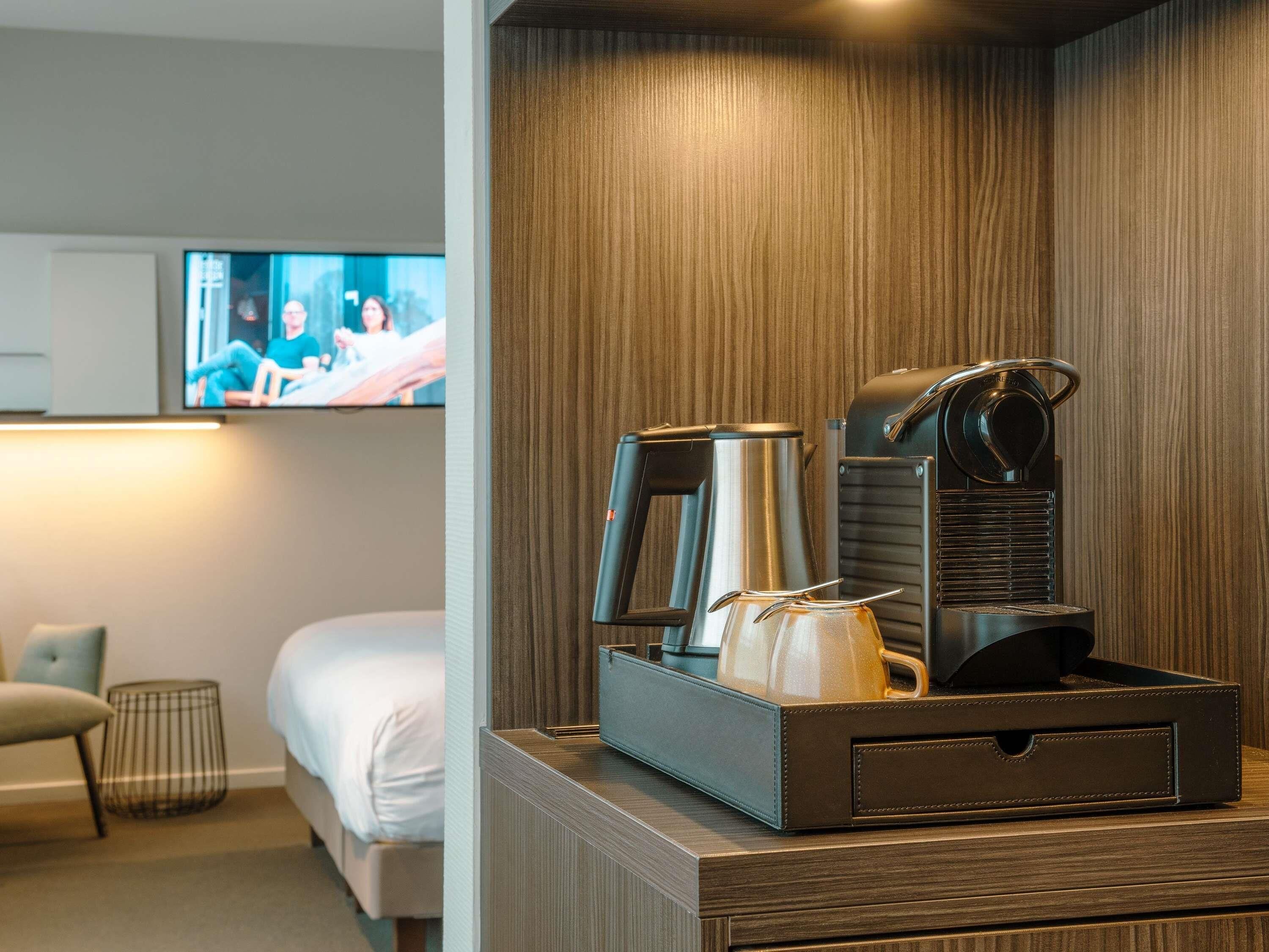 A hotel room with a bed, a TV mounted on the wall, a coffee maker, and a tray with a teapot and cups.