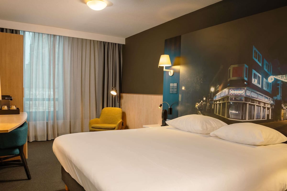 Een hotelkamer met een bed, een bureau met stoel, een lamp en een muurschildering.
