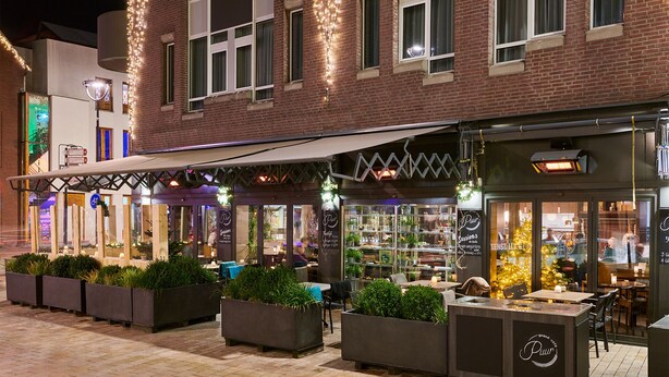 Ein Außenbereich mit Pflanzkübeln und Tischen, eine überdachte Terrasse und eine Schaufensterfront mit Glas.