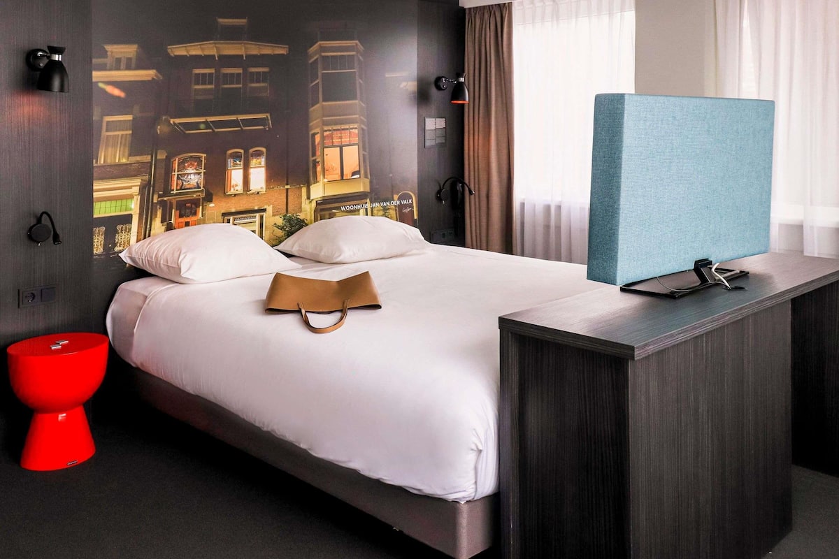 Een hotelkamer met een bed, een rode emmer, een blauwe scheidingswand en een muurschildering.